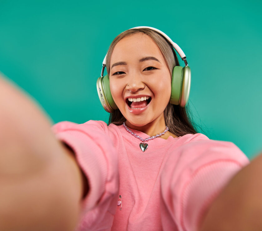 jolly and young asian woman in wireless headphones listening music and taking selfie on turquoise